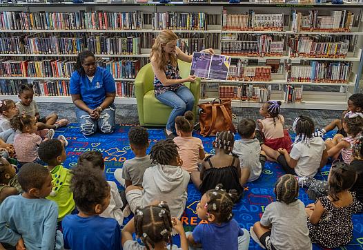 A lady sitting on a couch reads a book to kids sitting on the ground