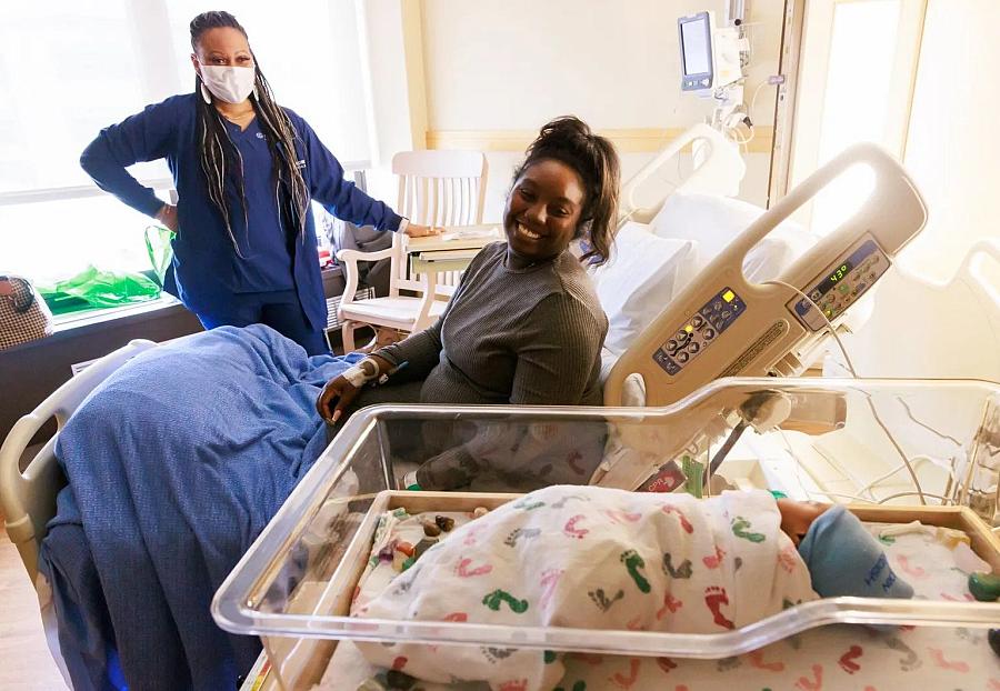 Woman on hospital bed watching her baby and health care worker in the mask is in the background.