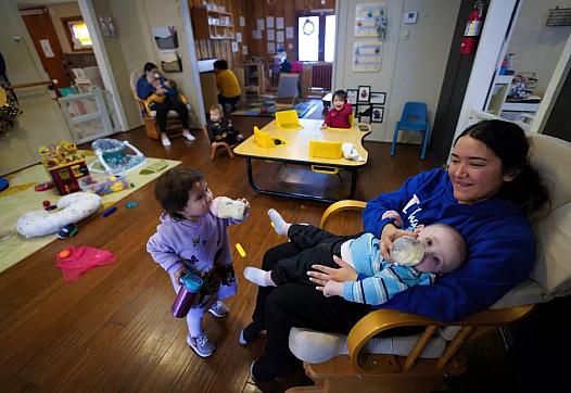 A girl feeding a baby and another child in front of her drinking milk from sippy cup